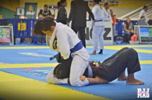 Dois lutadores de Jiu Jitsu executando um armlock em um dojo, com um lutador em kimono azul no chão e outro em kimono branco aplicando a técnica.