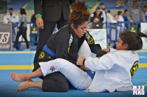 Dois lutadores de Jiu Jitsu no tatame praticando uma chave de braço, com garrafa de água, toalha e cronômetro na parede marcando 5 minutos.