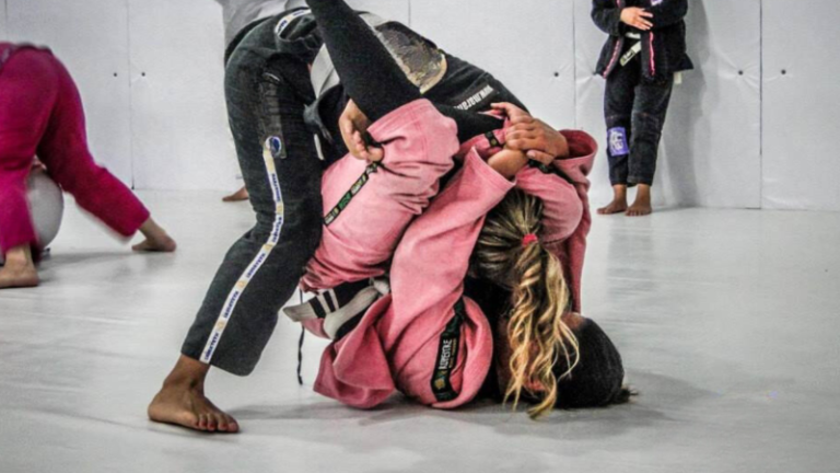 Dois praticantes de Jiu Jitsu no tatame, um aplicando o golpe do triângulo com as pernas em torno do pescoço do outro, ambos usando kimonos brancos com faixas pretas.