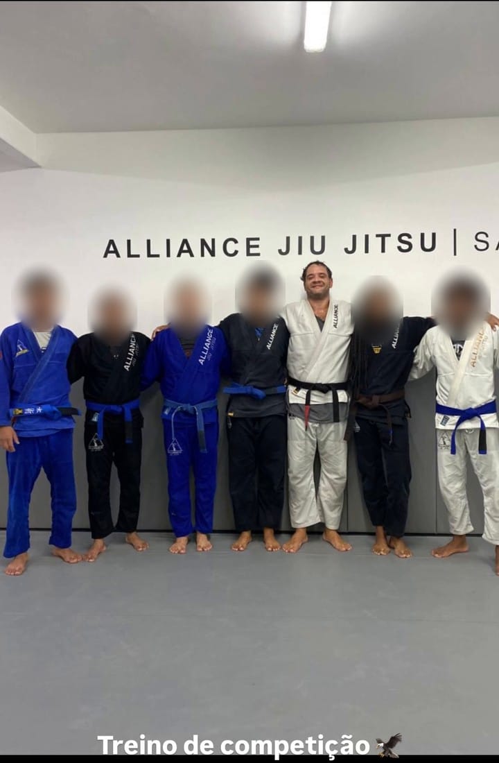 Rudá Boaventura de Macedo, de kimono branco e faixa preta, no tatame da Alliance Salvador, ao lado de outros praticantes com os rostos borrados.