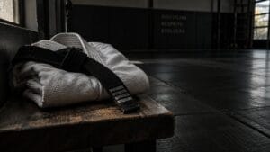 Kimono branco dobrado com faixa preta sobre um banco de madeira, numa academia de jiu-jitsu vazia e com pouca luz